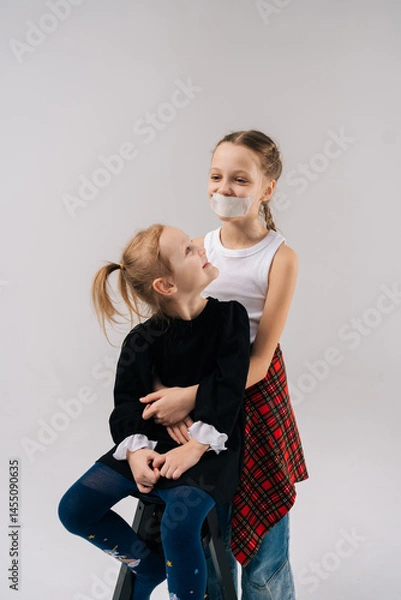 Obraz Compelling studio portrait showing young sisters hugging closely, one sibling wearing tape across mouth, symbolizing silenced voices on isolated white background. Concept of happy sisterhood.