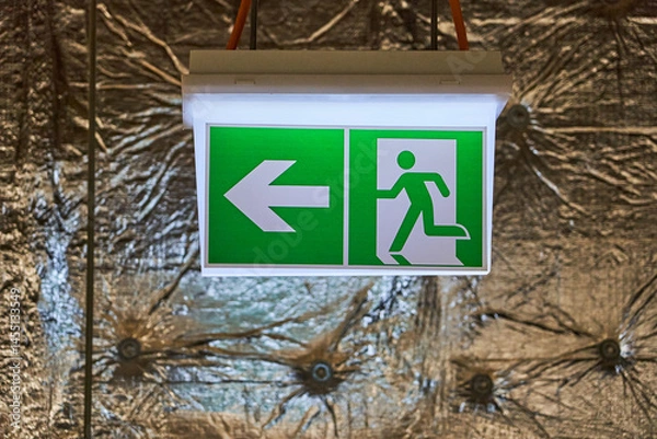 Obraz Illuminated Green and White Emergency Exit Sign with Arrow Pointing Left and Running Man Pictogram Mounted on a Wall