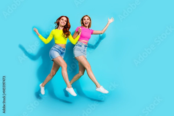 Obraz Young fashionable friends leaping with joy in vibrant summer outfits against a teal background, embodying friendship and youthfulness.
