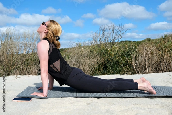 Obraz Women practicing yoga outdoors on sandy surface, performing upward facing dog pose on mat, enjoying sunny weather, clear sky, and natural surroundings for relaxation and wellness
