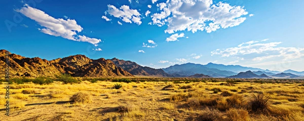 Fototapeta Experience the vast beauty of a desert landscape, featuring golden sands, rugged mountain ranges, and a clear blue sky with fluffy clouds, capturing the essence of nature's tranquility.