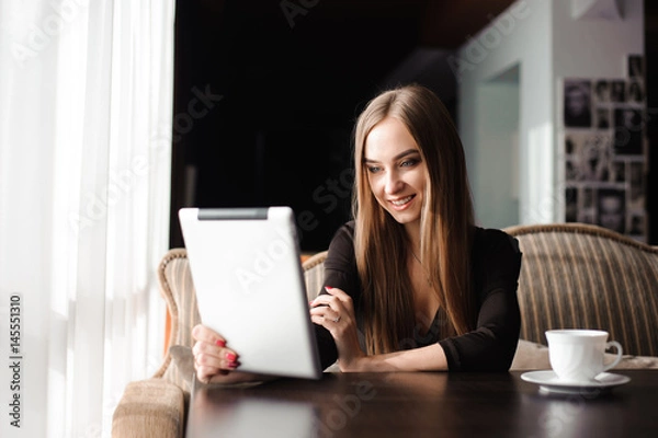 Fototapeta Happy young woman drinking coffee / tea and using tablet computer in a coffee shop
