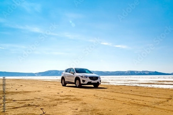 Obraz A white car in the spring on a sandy beach in front of a river, mountains, melting ice