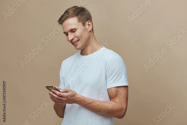 Fototapeta Smiling young man in casual white t-shirt using mobile phone, cheerful expression, engaging with device, casual indoor setting, beige background, lifestyle, everyday life, modern technology concept