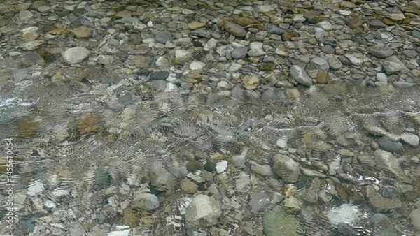 Fototapeta Clear water flows gently over smooth stones in a serene riverbed. Sunlight glimmers on the surface, creating a peaceful atmosphere in nature