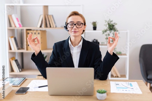 Obraz Adult woman meditating at office desk in lotus pose wearing glasses and suit promoting work-life balance and mindfulness during break period enhancing productivity and maintaining demeanor.