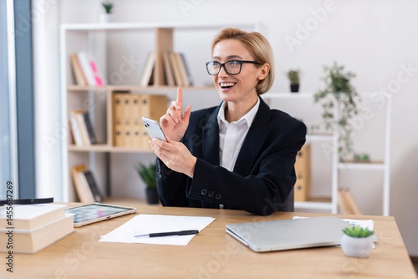 Obraz Smartly dressed businesswoman seated at desk using smartphone with happy expression. Office features organized bookshelf, papers, and plants creating professional environment.