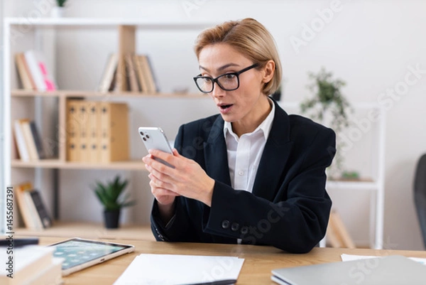 Obraz Professional adult Caucasian woman wearing formal suit sitting at desk expressing surprise while looking at smartphone screen in modern office with organized shelves in background.