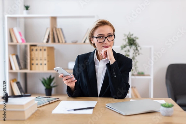 Obraz Businesswoman in formal attire working at desk holding smartphone surrounded by documents and a laptop. Professional environment with organized shelves and plants adding comfort workspace.