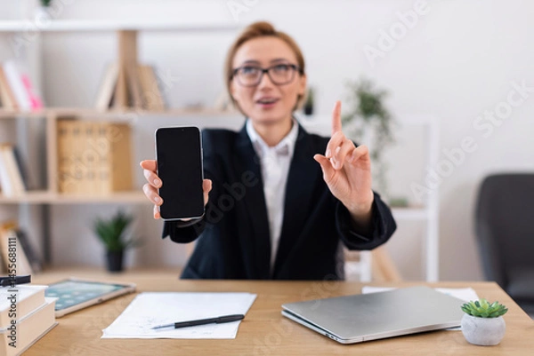 Obraz Businesswoman with glasses pointing while holding smartphone during professional discussion at office. Image portrays modern work, technology importance, communication in business environment.