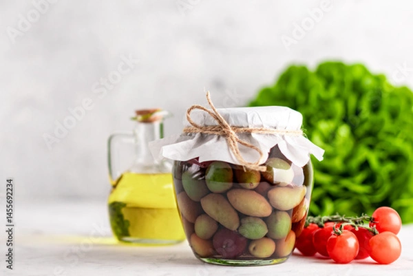 Fototapeta A glass jar with various pickled olives in brine stands on a white table.