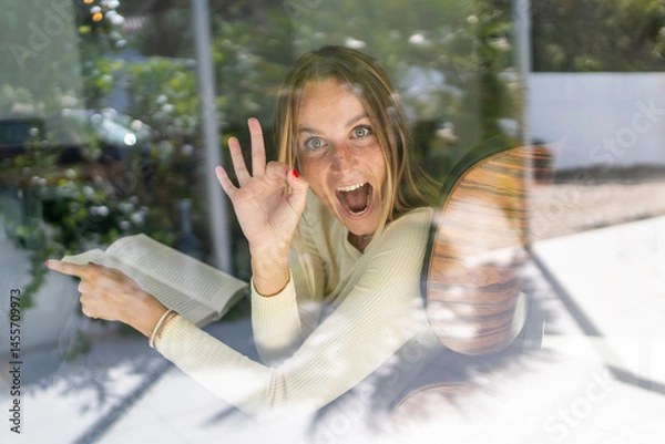 Fototapeta Happy playfull woman by the window at home, doing OK gesture and smiling with a book on her hands