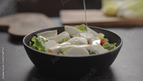Obraz pour walnut oil onto salad with romaine lettuce, tomatoes and mozzarella in black bowl on concrete countertop