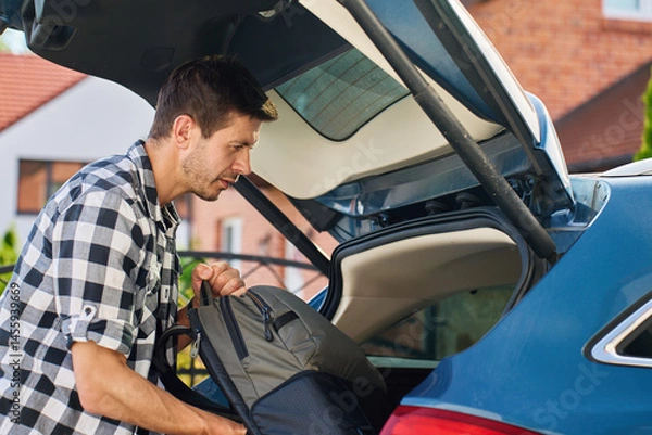 Obraz Man loading backpack into car trunk for road trip. Preparing for travel by packing luggage into vehicle. Outdoor travel concept