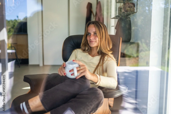 Obraz Happy young woman by the window at home, starting new day, enjoying wellbeing with tea or coffee mug on her hand.