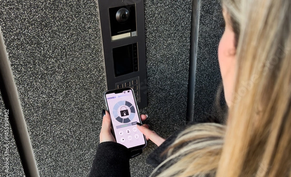Fototapeta Young stylish woman getting access to the building by attaching smartphone to intercom. Concept of modern security technologies for access and smart home