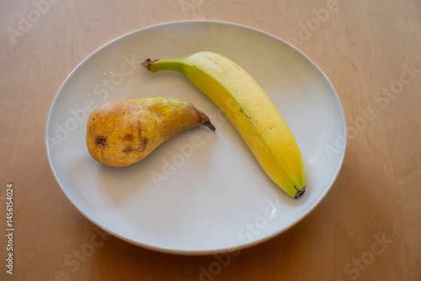 Fototapeta One pear and one banana in a white dish on a wooden table