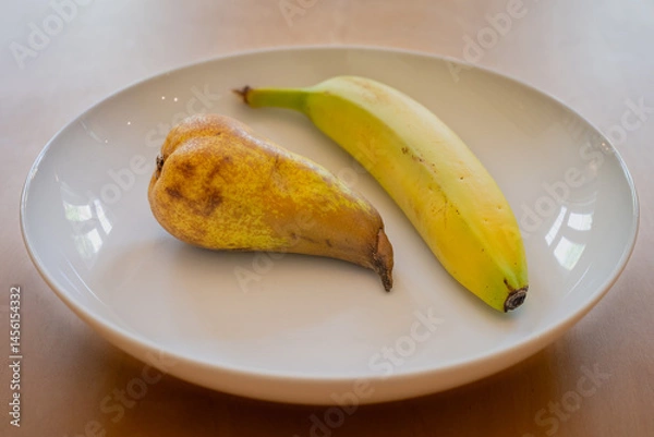 Fototapeta One pear and one banana in a white dish on a wooden table
