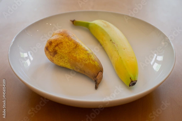 Fototapeta One pear and one banana in a white dish on a wooden table