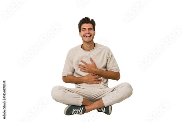 Fototapeta Young caucasian man sitting on the floor isolated on white background laughs happily and has fun keeping hands on stomach.
