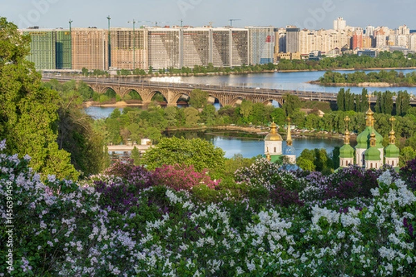 Fototapeta Scenic spring view of Kyiv with blooming lilacs, golden domes of Vydubychi Monastery, the Dnipro River, and modern residential buildings on the city's left bank.