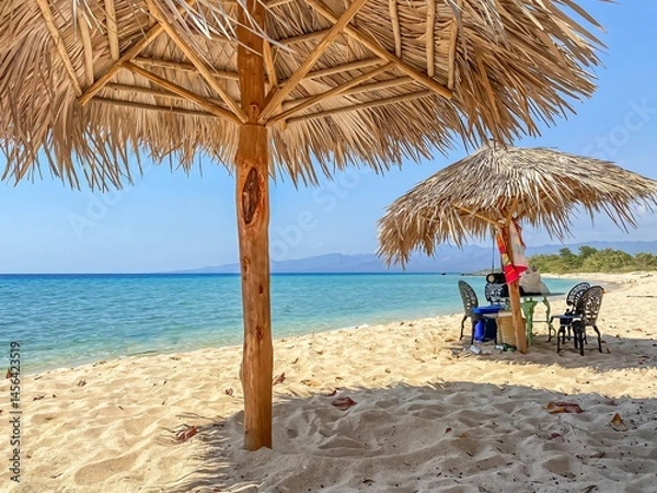 Fototapeta Straw beach umbrellas providing shade on a beautiful white sand beach in cuba, with turquoise water and mountains in the background