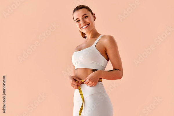 Fototapeta Smiling fit young woman in white sportswear measuring her waist with a tape measure on a soft peach background. Concept of healthy lifestyle, fitness tracking, and workout motivation.