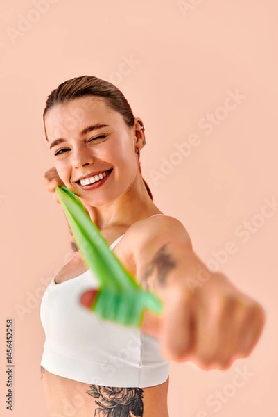 Fototapeta Portrait of a young woman in a vertical image, pointing a resistance band toward the camera while smiling and playfully winking.