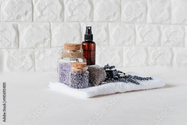 Fototapeta Lavender Spa Set with Dried Flowers and Bath Salts. Relaxing Spa Arrangement. Lavender flowers, bath salts, dried buds in glass jars, spray bottle, and sachet on white towel