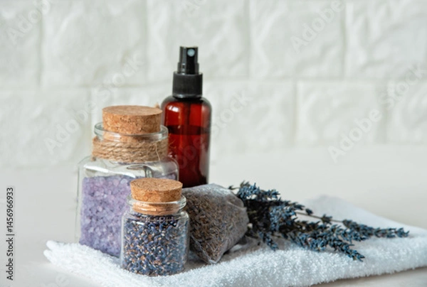 Fototapeta Lavender Spa Set with Dried Flowers and Bath Salts. Relaxing Spa Arrangement. Lavender flowers, bath salts, dried buds in glass jars, spray bottle, and sachet on white towel