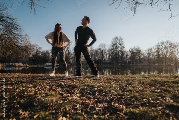 Fototapeta A couple stands together in a park during sunset, enjoying the serene scenery and shared moment by a peaceful lake with trees in the distance.