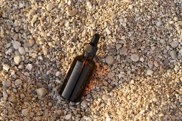Fototapeta Close-up of a brown mockup of a glass jar of serum with a dropper on a small pebble on the beach. Concept of natural cosmetics for face care, wrinkle reduction, moisturizing. Image for your design