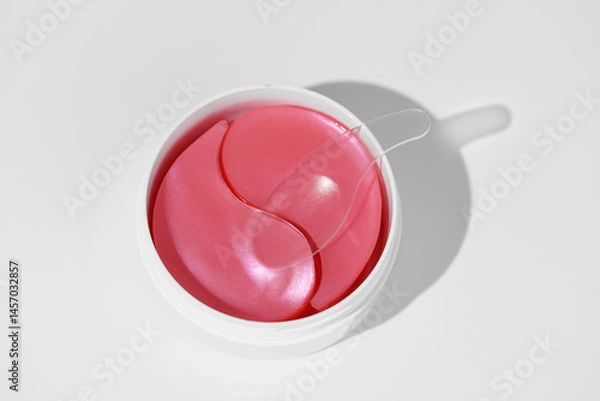 Fototapeta White skincare jar with pink hydrogel eye patches and a spatula on white blank background. Reduces swelling, brightens dark circles, and smoothes delicate under eye skin concept.