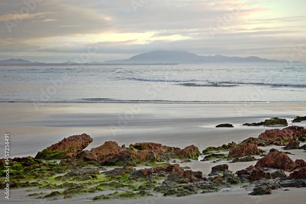 Obraz Red rocks covered by green moss on the beach sand,  waves breaking on water edge, foggy morning with cloudy sky in Itacuruca beach, Cananeia, Sao Paulo state