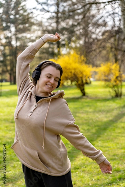 Fototapeta Smiling woman stretching with headphones on, enjoying music in a spring park. Morning routine, stretching, music therapy.