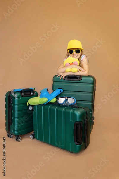 Fototapeta Cheerful little girl in yellow glasses and hat with toy duckling stands behind green travel suitcases with flippers and diving mask on them. Family on vacation. Sea travel concept.