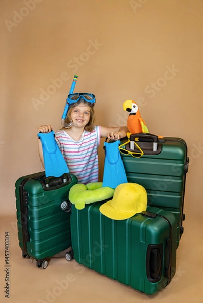 Fototapeta Little girl in diving goggles with flippers in hands and parrot toy next to her sits near green travel suitcases, ready for travel. Child on vacation or holiday. Travel concept.
