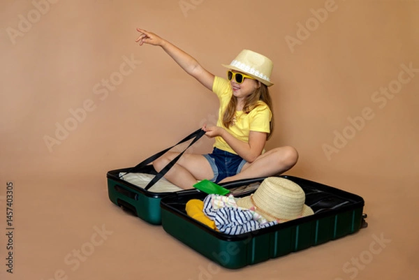 Fototapeta Little girl in yellow glasses and hat sitting in suitcase and pointing direction with cuckoo ready for travel. Background with space for text. Child on vacation or holiday. Travel concept.