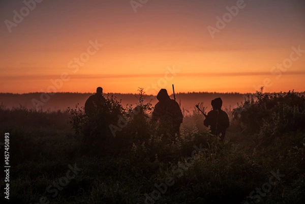 Fototapeta Group of hunters with shotguns going through rural field towards forest during sunset with orange sky and forest on horizon 