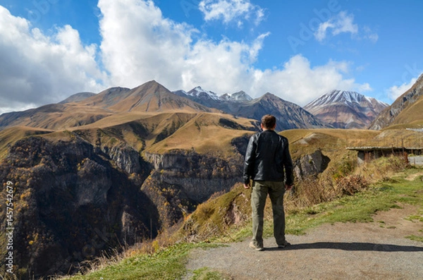 Fototapeta Man enjoys an incredible Caucasus Mountains view featuring valleys and peaks, basking under a clear blue sky. The scene exudes adventure, freedom, and the dominance of nature's breathtaking beauty
