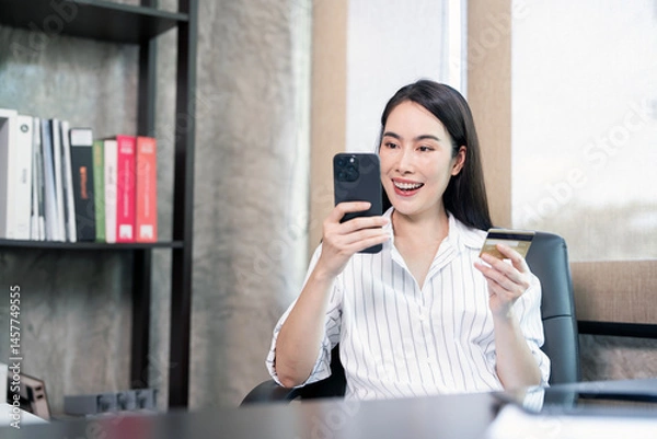 Fototapeta A young woman sitting at a desk happily holding a smartphone and credit card, likely shopping online or making a mobile payment in a modern office setting.