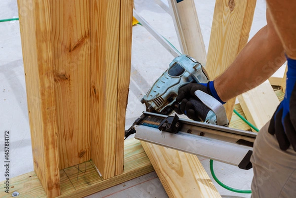 Obraz Construction worker skillfully operates nail gun, securing wooden beams to frame structure at busy works site.
