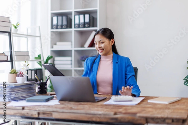 Fototapeta A focused asian woman accountant analyzing paperwork while calculating expenses using laptop and calculator at her office desk.
