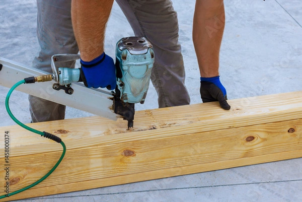 Obraz Worker drives nails into wooden beam using air nail gun on construction site under works day
