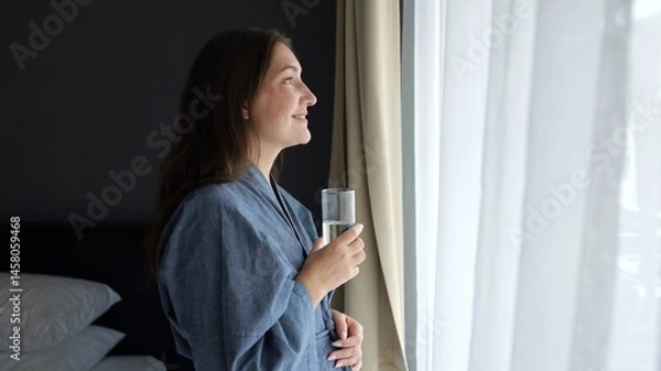 Fototapeta Relaxed woman drinks fresh water by window smiling slightly