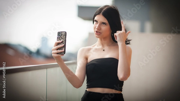 Obraz A trendy young girl stands on a balcony, capturing a selfie with her smartphone. Her confident pose highlights her modern style and fashionable outfit, perfect for social media