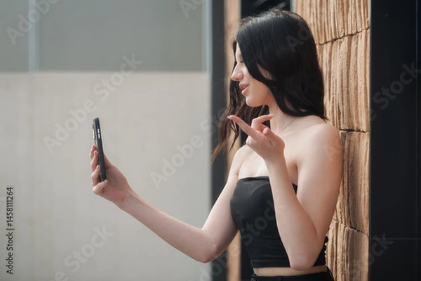 Obraz A fashionable young girl is taking a selfie in a stylish outfit against a contemporary textured wall. She strikes a playful pose, capturing the essence of social media influence during the day