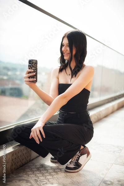 Obraz A fashionable young girl is sitting on a stylish ledge, smiling as she captures a selfie with her phone. The background shows beautiful urban scenery, indicating a relaxing afternoon