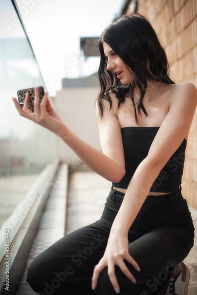 Obraz Young social media influencer captures a stylish selfie on a balcony. Dressed in a chic black outfit, she showcases her modern fashion sense while enjoying the sunny day