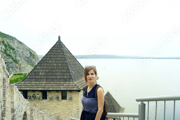 Fototapeta Exploring the Golubac fortress: a smiling woman stands on the defensive wall against the backdrop of tower number 4, part of the blue tourist route. A female tourist during a trip to Serbia.